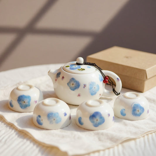 Luminous white porcelain tea set with real pressed blue hydrangea relief on a linen surface, gift box in soft natural light.