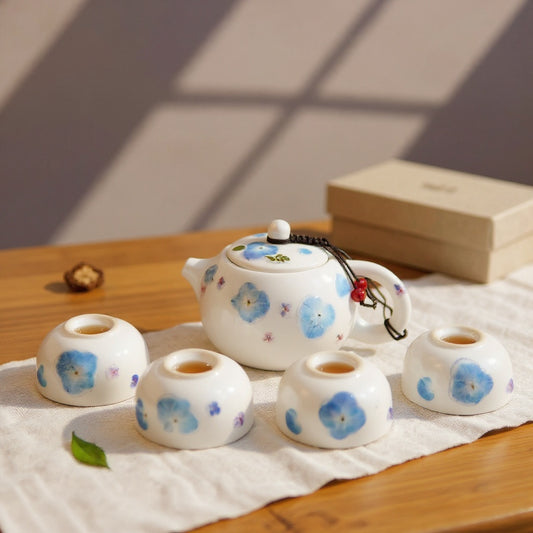 Zen tea set with white porcelain teapot and four teacups featuring real pressed blue hydrangea petals on a natural wood surface, gift box in background under soft daylight