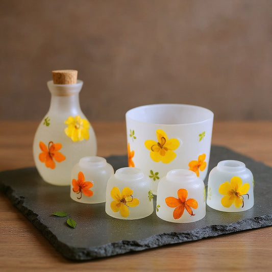 A 6-piece frosted glass sake set with embedded yellow pressed flowers arranged on a slate surface in soft natural light, wabi-sabi styling.