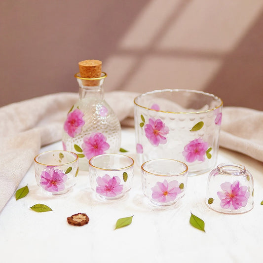 Hero image of a 5-piece frosted glass sake set with embedded peach blossoms and gold rims on a marble surface with botanical accents.