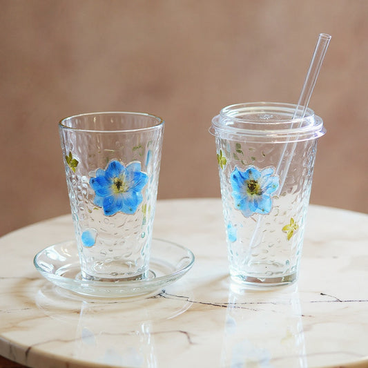 Handcrafted glass cup with real pressed botanical relief on marble surface with soft natural light, accompanied by saucer, lid, and straw.
