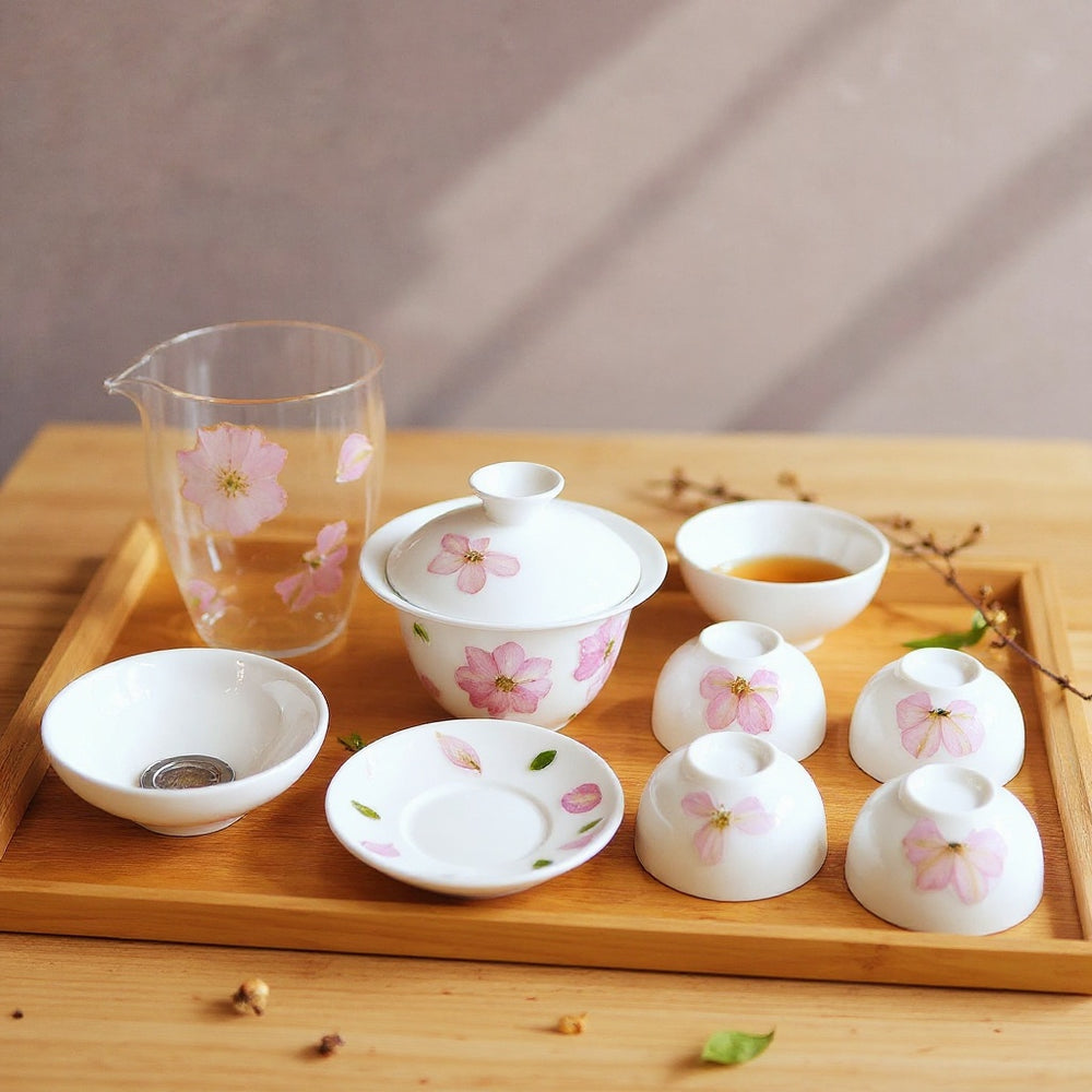 Luminous white porcelain cherry blossom gaiwan and teacups on a natural wood surface with fresh cherry blossom sprigs, soft daylight, minimalist Japanese-inspired composition.