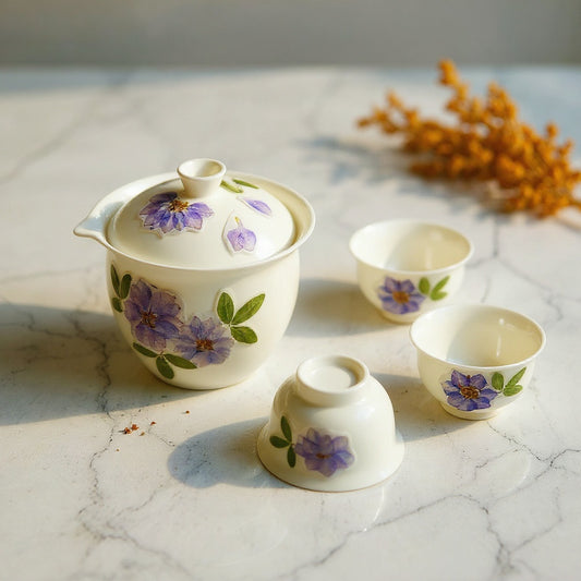 Semi-translucent white porcelain gaiwan and teacups with embedded dried flowers on marble amid botanical props.