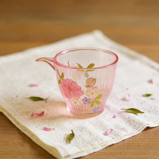 Hand-blown gradient pink glass cup with preserved flowers and butterfly motif on linen with dried petals, wide editorial shot