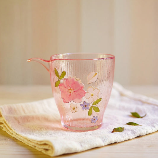 A hand-blown gradient pink glass cup with raised preserved flowers and a butterfly motif, resting on a linen cloth with scattered dried petals, in soft natural lighting.