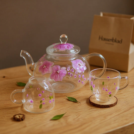 Hand-blown glass teapot and cups with real pressed flower petals embedded in raised relief, displayed on wood with kraft packaging in background.