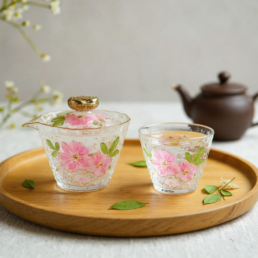 A delicate glass gaiwan and fair cup set with real dried flowers embedded under resin on a wooden tea tray, surrounded by loose dried flowers and a small teapot, soft natural light.