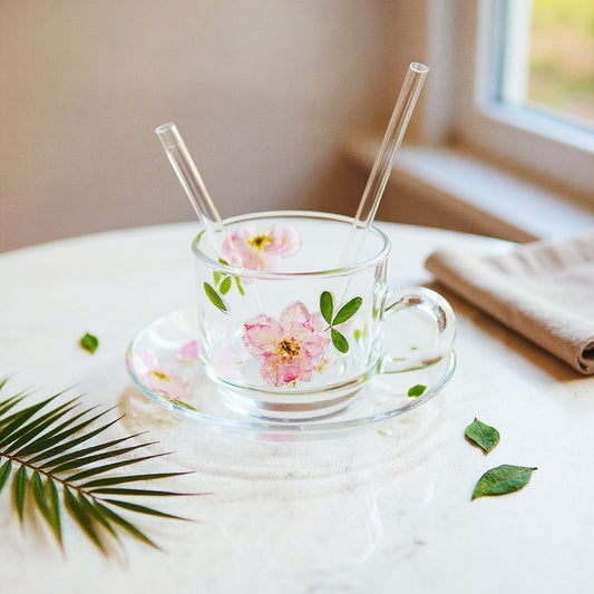 Elegant pressed flower glass cup set with saucer, straw, and lid, real embedded pink petals and green leaves, on white marble with natural accents.