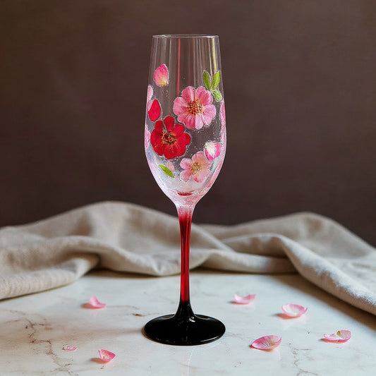 A tall elegant champagne flute with vivid red stem and matte black base, its bowl adorned with raised soft pink preserved flower petals and green leaves, rests on a marble surface with gentle side lighting and negative space.