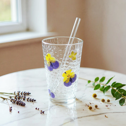 Delicate clear glass tumbler with embedded real dried flower petals and green leaves, matte lid and glass straw, on marble surface with dried botanical accents, soft natural lighting.