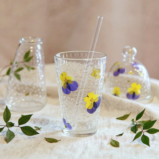A handmade pressed flower glass cup with straw on a linen cloth, surrounded by dried botanicals, soft lighting.