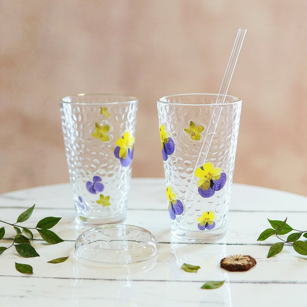Clear glass tumbler with raised real dried flower petals and green leaves, matte lid and glass straw, on marble surface with botanical decor, soft natural light.