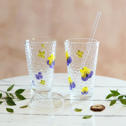 Clear glass tumbler with raised real dried flower petals and green leaves, matte lid and glass straw, on marble surface with botanical decor, soft natural light.