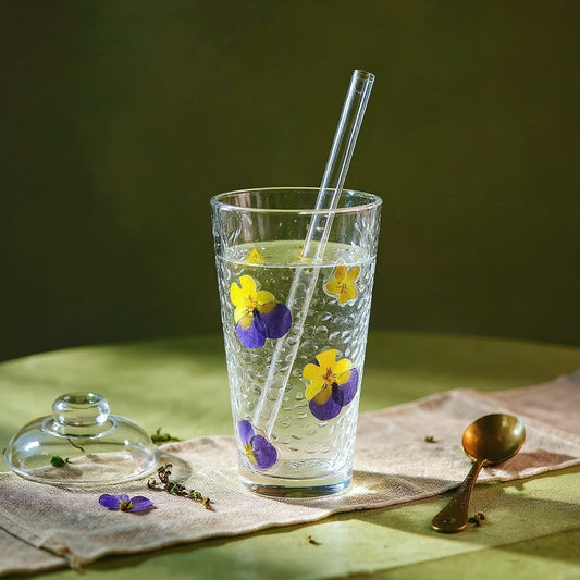 Clear glass tumbler with real pressed flowers and leaves, lid and straw, on linen with dried botanicals against a sage and amber gradient background.
