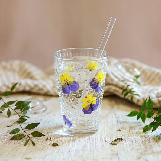 Hero image of a vintage European style floral glass tumbler with raised dried flower petals and green leaves, matte lid, and clear straw, styled on a linen surface with botanical props in soft natural light.