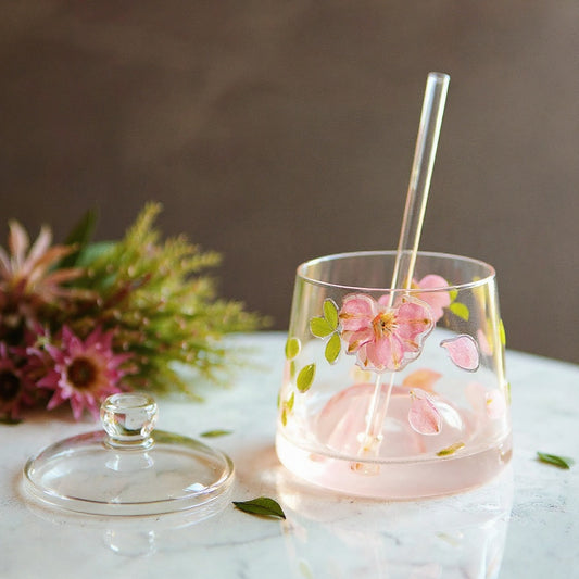 A clear stemless glass cup with pink dried flowers and green leaves embedded in raised relief, paired with a glass lid and straw, set on a marble surface with fresh botanicals.
