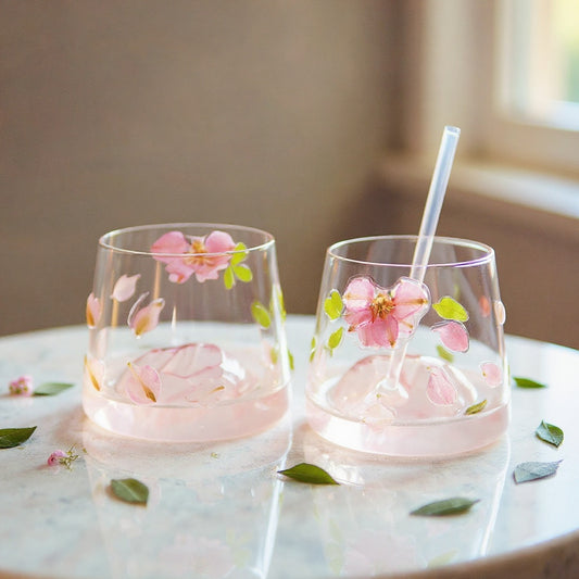 Stemless glass cup with embedded real pink petals and green leaves, glass lid and straw, styled on marble with fresh botanicals, soft natural light.