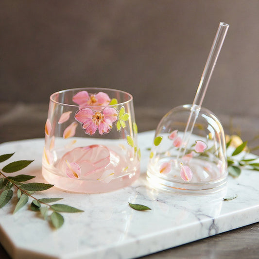 Clear stemless glass cup with embedded pink petals and green leaves, with matching lid and glass straw, styled on marble with fresh botanicals, soft natural lighting.